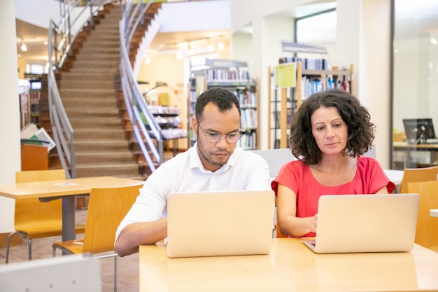 College students collaborating on laptops in a bright, modern library. They are surrounded by open books and notebooks, suggesting a scene of focused studying and research, highlighting the resources available at college.